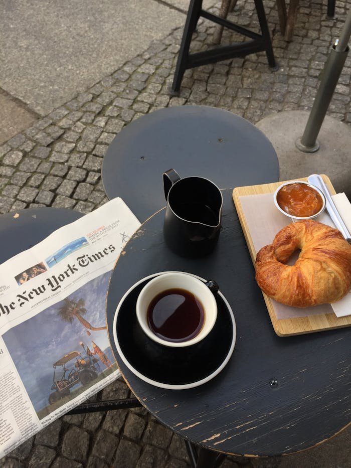 A cozy setup in Berlin featuring coffee, croissant, jam, and a newspaper on a cafe table.