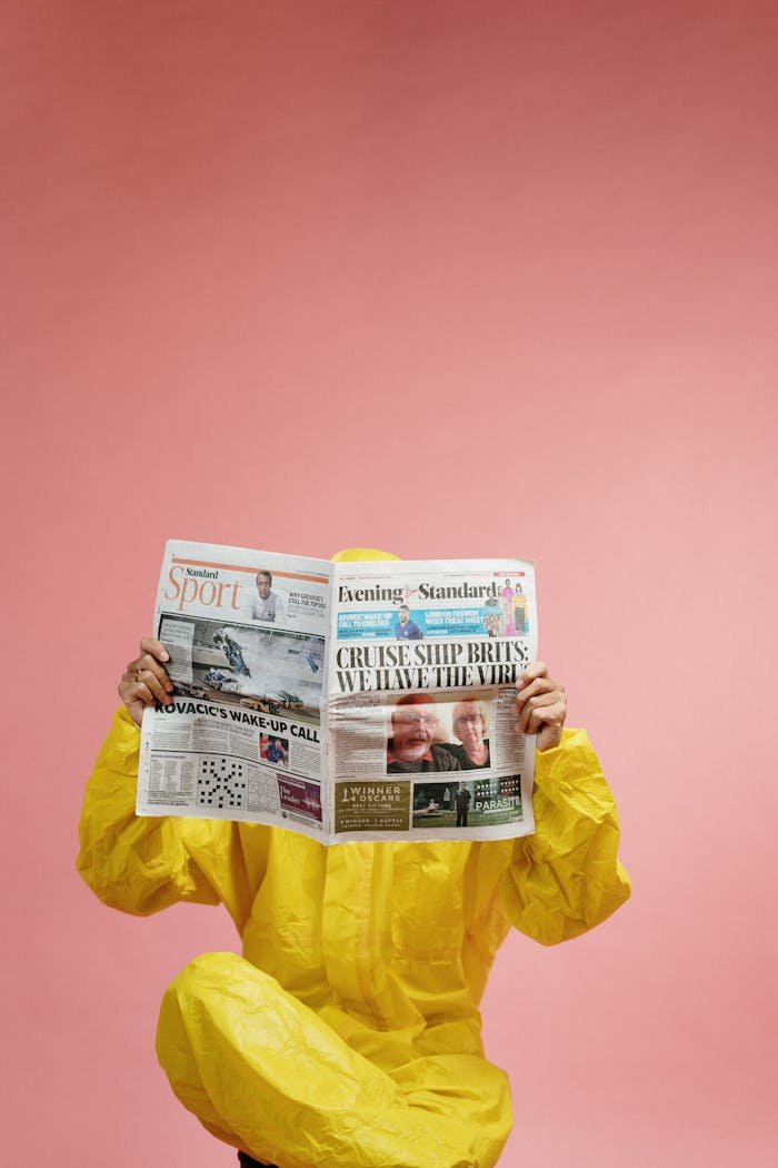 Individual in protective suit reads newspaper with pandemic headline against pink backdrop, symbolizing COVID-19 era.